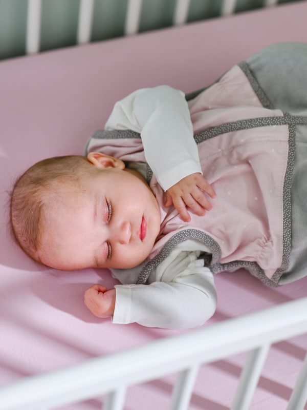 A baby in a pink LEN sleep bag on a pink LEN fitted sheet is asleep in a white GULLIVER cot.