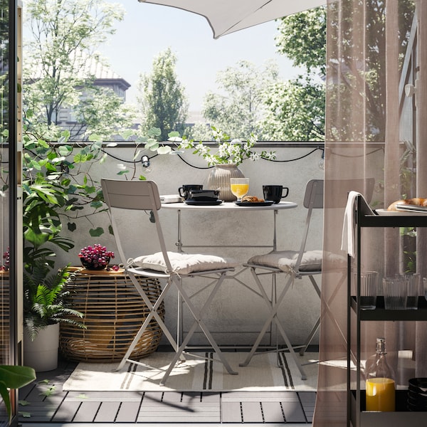 A balcony with a grey SUNDSÖ outdoor table and two grey outdoor chairs on a rug, plus a coffee table, plants and a parasol.
