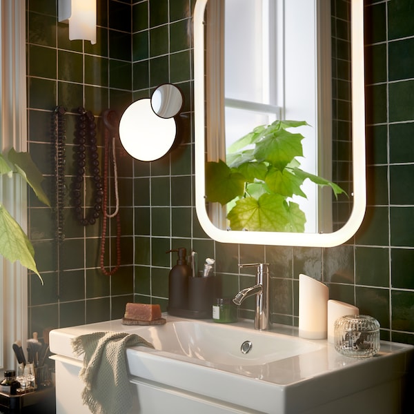 A bathroom with a white mirror with integrated lighting over a wash-stand, plus two LED block candles and a potted plant.