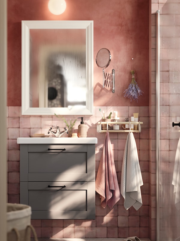 A bathroom with pink tiles has a grey ENHET washstand and a white mirror next to a shelf with two towels on hooks.