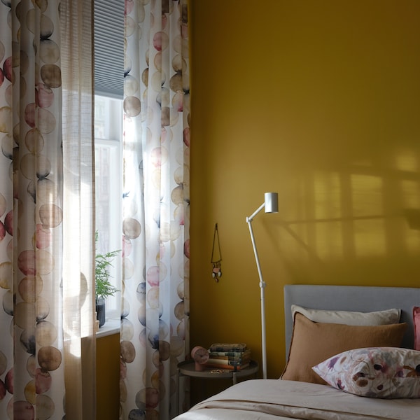 A bed and a white floor/reading lamp by a window with multicolour SÄCKMAL curtains and a block-out cellular blind.