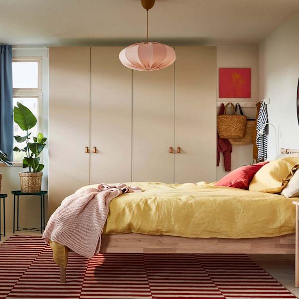 A bedroom with a bed frame and two chests of drawers in birch, a gray-beige wardrobe, blue curtains and yellow bed textiles.