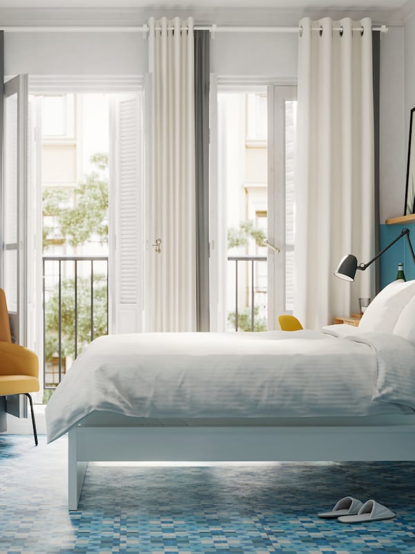 A bedroom with a white bed with white bed linen, blue walls and floors and French windows opening onto a balcony.