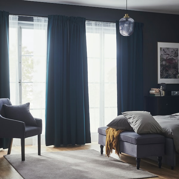 A bedroom with dark turquoise MAJGULL block-out curtains and net curtains in large windows near a bed and a bench.