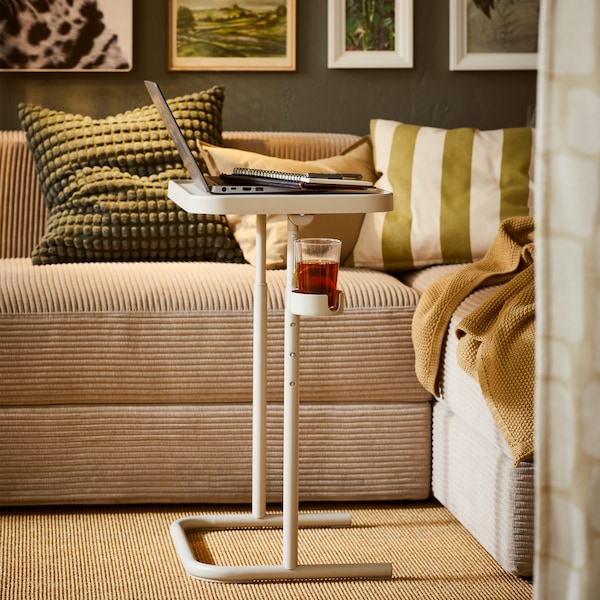 A beige BJÖRKÅSEN laptop stand by a grey-beige JÄTTEBO sofa dotted with textiles. A row of framed art hang above the sofa.