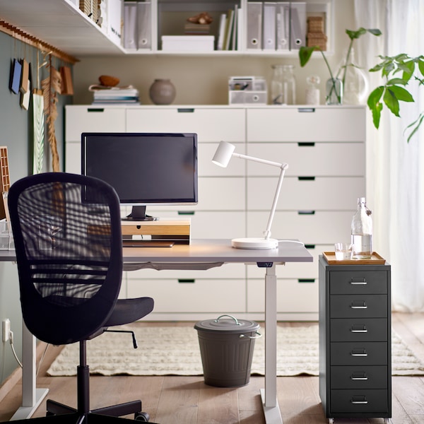 A black office chair and a HELMER drawer unit by a grey/white RODULF sit/stand desk with a work lamp and computer screen.