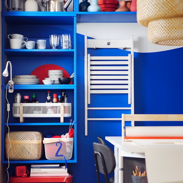 A blue PLATSA open shelving unit stores several baskets and a white FRÖSVI folding chair is hanging on hooks next to it.