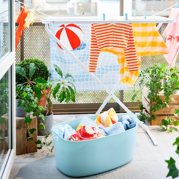 A blue TORKIS flexi laundry basket filled with laundry and a white drying rack on a sunny balcony decorated with plants.