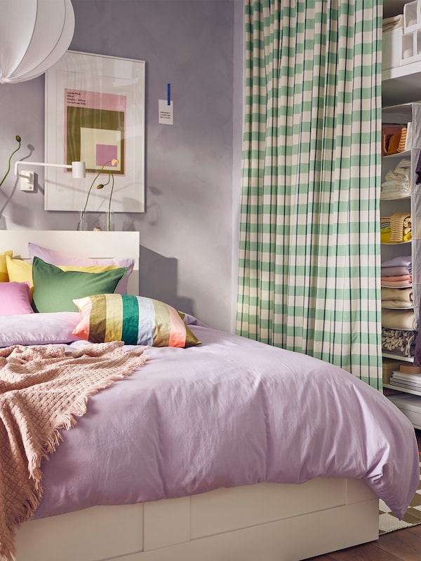 A BRIMNES bed in a lilac room covered in lilac NATTSVÄRMARE bed linen. A green GLANSHAGTORN curtain covers clothing storage.