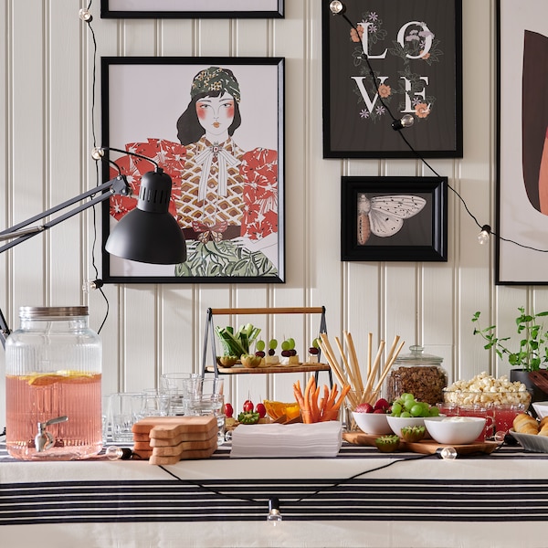 A buffet table with food and drink in various bowls, dishes and a VARDAGEN jar with tap, by a wall with pictures in frames.