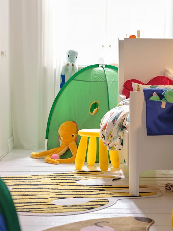 A BUSUNGE extendable bed on one side of a small bedroom with a yellow MAMMUT stool by it and a green play tent behind it.