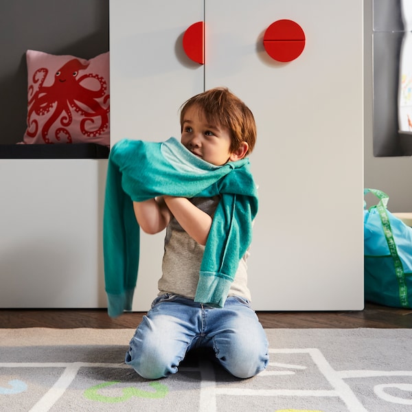 A child getting dressed on a grey rug in front of a white bench with toy storage and a white wardrobe with a pull-out unit.