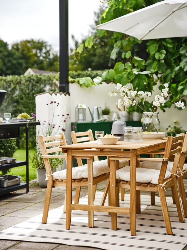 A corner of a patio surrounded by vegetation with an ASKHOLMEN table and four ASKHOLMEN chairs with armrests on a rug.