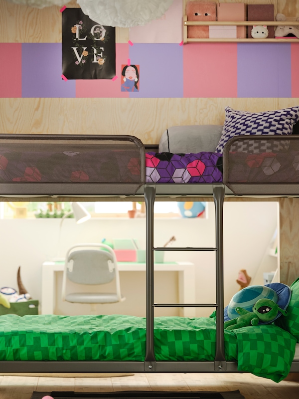 A dark grey loft bed with a ladder, purple and pink storage cabinets on the wall above it and a white desk with a chair.