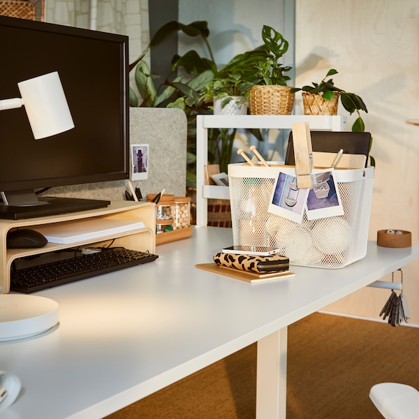 A dimly sunlit workspace with a TROTTEN sit-stand desk, a VATTENKAR laptop stand and a RISATORP basket holding accessories.