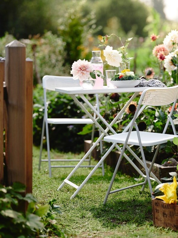 A flowery outdoor garden features two TORPARÖ chairs and a TORPARÖ table with pink drinks, vases and a salad dish on it.