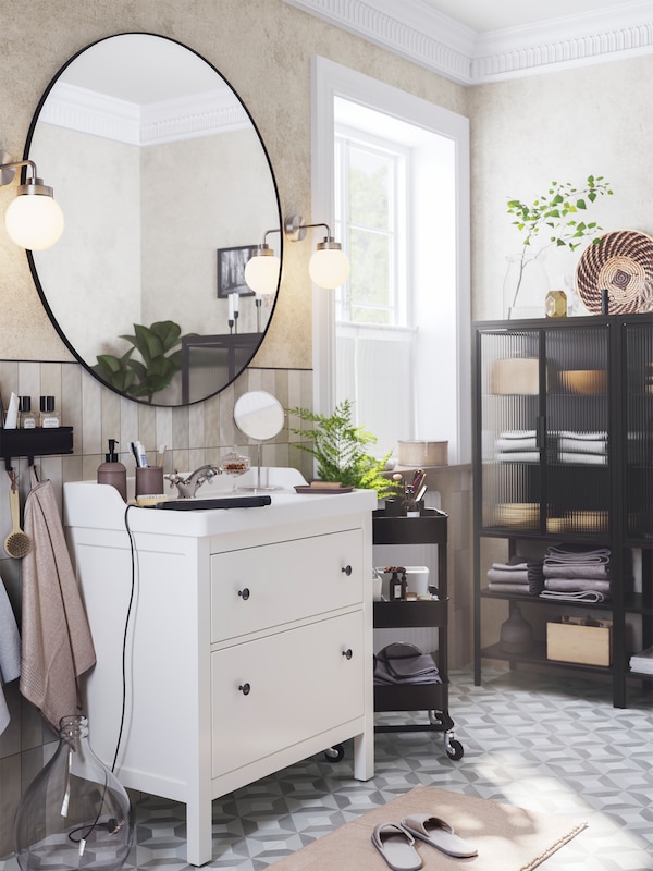 A grey bathroom with a white HEMNES sink cabinet has a large mirror, a black RÅSKOG trolley and a MOSSJÖN glass door cabinet.