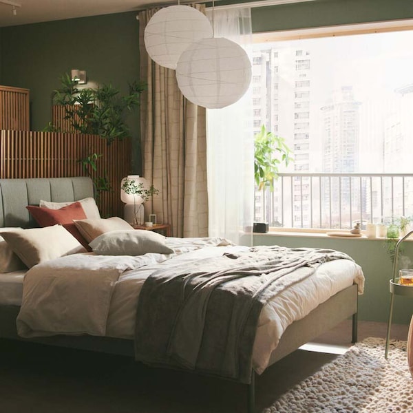 A grey-green TÄLLÅSEN upholstered bed frame with privacy screens behind it is in a serene green bedroom with large windows.