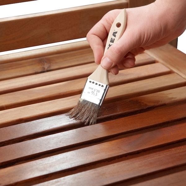 A hand holding a paint brush applying VÅRDA wood stain to the surface of a piece of outdoor furniture.