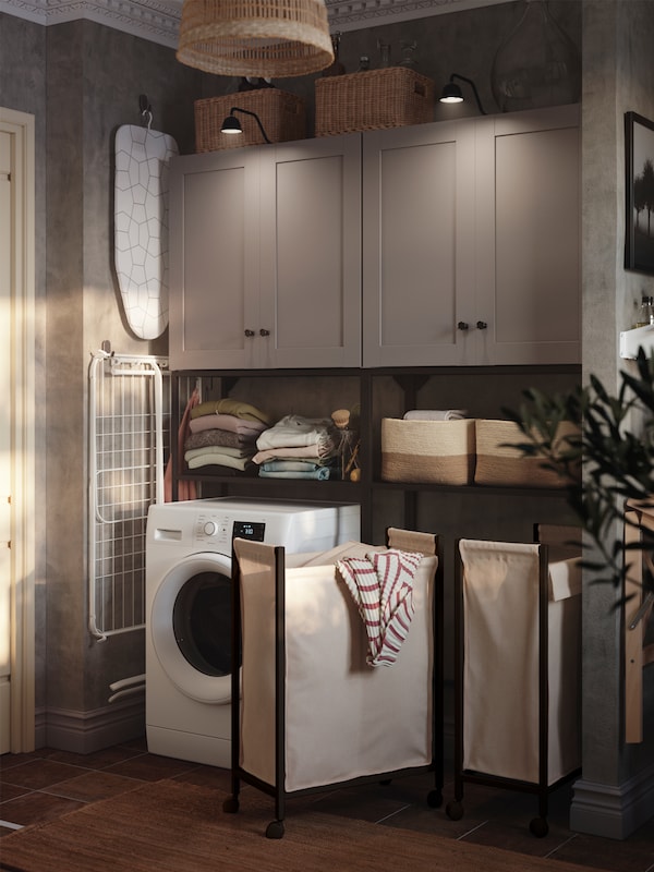 A laundry room has an ENHET storage combination with baskets on shelves, a washing machine and two laundry bags on castors.