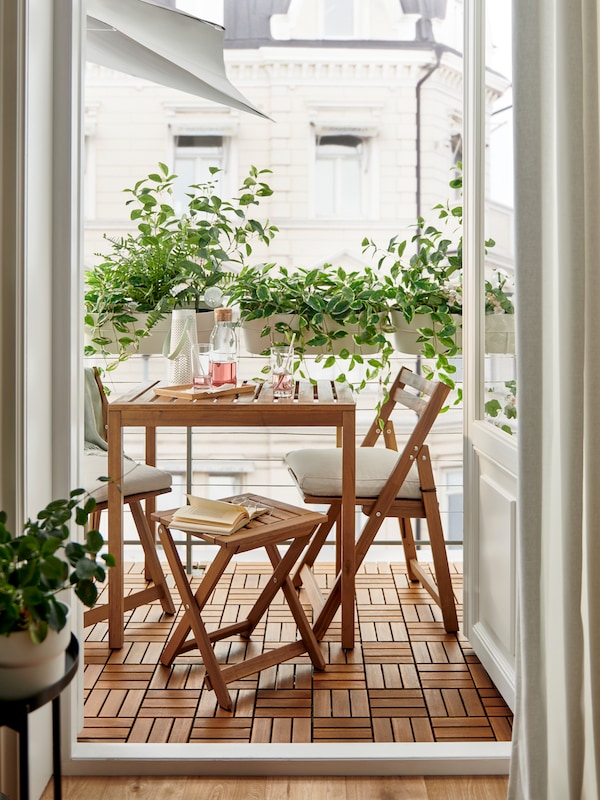 A light-brown-stained NÄMMARÖ outdoor table, a foldable stool and foldable chairs on a balcony enveloped in leafy plants.