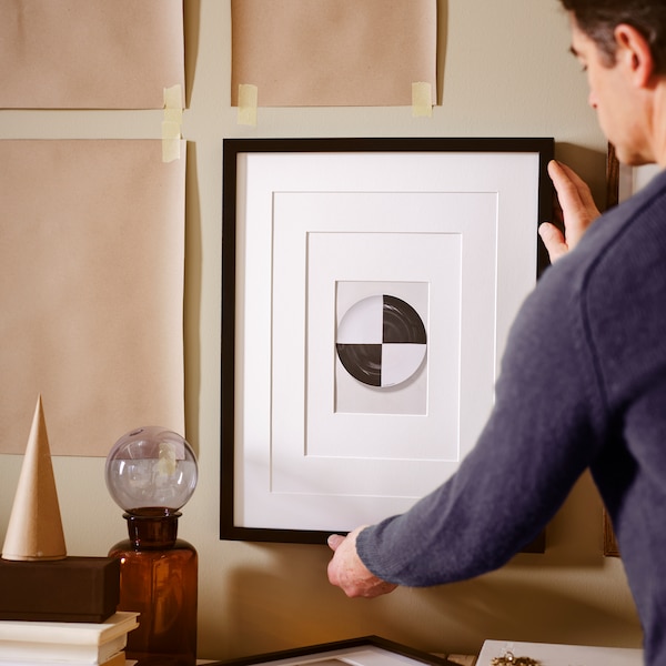 A man hanging a picture in a black RIBBA frame on a beige wall with brown paper templates taped to it.