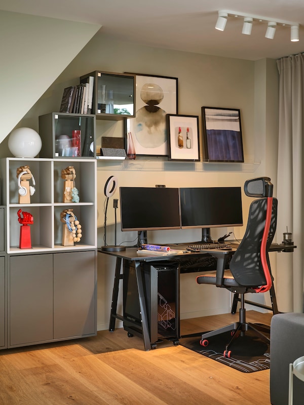 A MATCHSPEL gaming chair and UTESPELARE gaming desk in the corner of a grey room holding two monitors and other accessories.