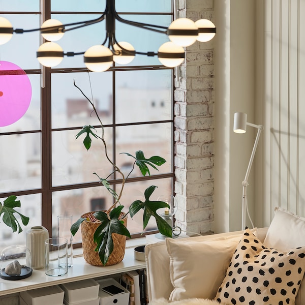 A NYMÅNE floor/reading lamp stands beside a sofa near a window. An 8-armed STORSLINGA LED chandelier hangs from the ceiling.