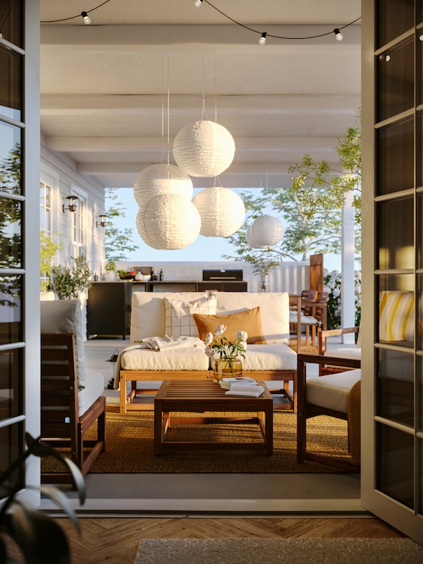 A patio seen from indoors featuring a NÄMMARÖ 4-seat conversation set under some SOLVINDEN LED solar-powered pendant lamps.