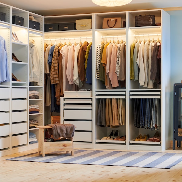 A PERJOHAN stool with storage stands in front of a large open PAX corner wardrobe with clothes, shoes, boxes and bags.