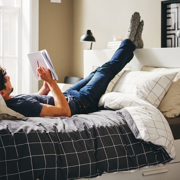 A person lies on a white BRIMNES bed with storage and headboard, with their feet on the headboard, holding a book and pencil.