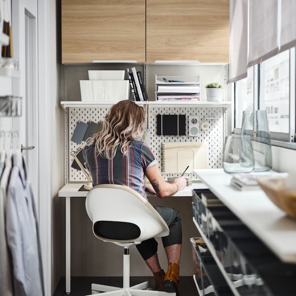 A person sitting in a beige/white swivel chair by a white BOAXEL/LAGKAPTEN shelving unit with table top on a balcony.