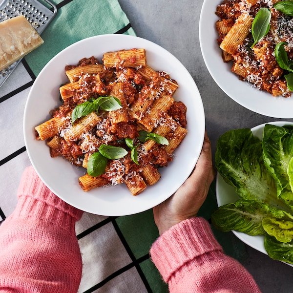 A person wearing a pink sweater is holding a plate containing plant-based Bolognese and pasta with a sprig of basil on top.