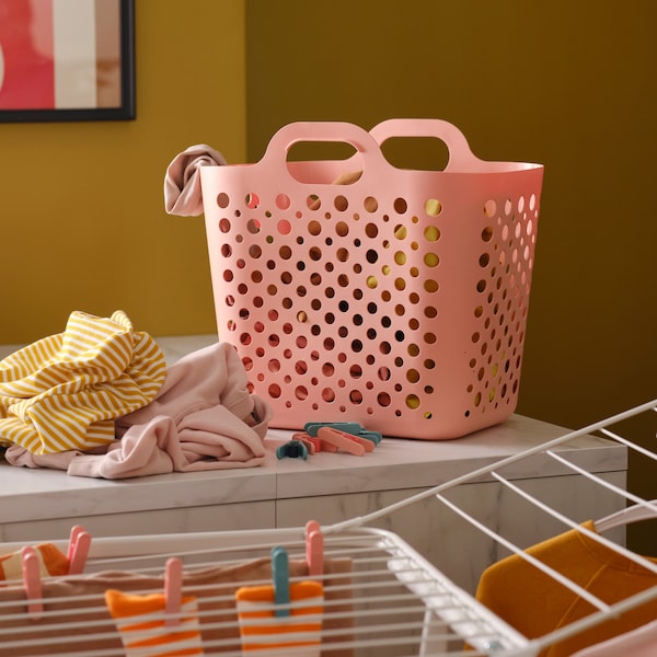 A pink SLIBB flexible laundry basket and a pile of clothes on a kitchen worktop, by a white drying rack with more clothes.