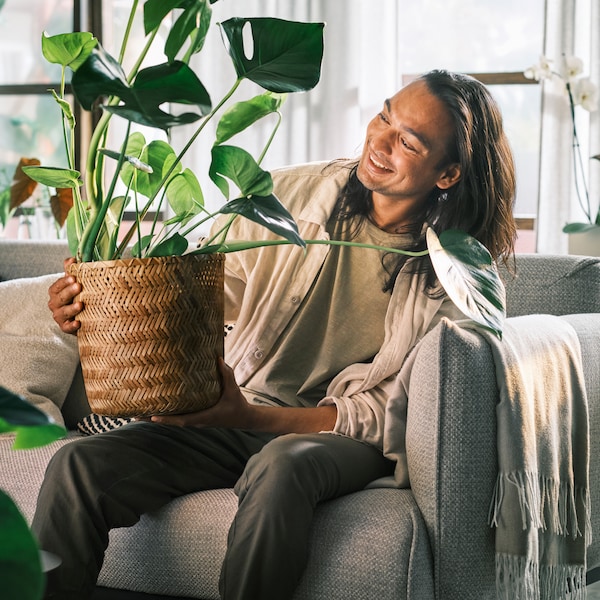 A smiling person sitting on a light-grey ÄPPLARYD three-seat sofa holding a monstera planted in a KLYNNON bamboo plant pot.