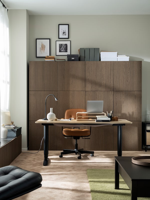 A softly sunlit room with a BEKANT sit-stand desk, a golden-brown ALEFJÄLL office chair and a wall of wood-front cabinets.