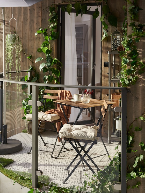 A sunny balcony features a TÄRNÖ table and two TÄRNÖ chairs with chair cushions on them, light grey floor decking and plants.