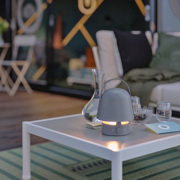 A VAPPEBY Bluetooth speaker lamp, with the light turned on, is placed on a SEGERÖN coffee table in an outdoor lounge area.