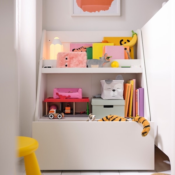 A white BERGIG book display with storage stands in a corner, holding a lamp and a lot of books, boxes and toys.
