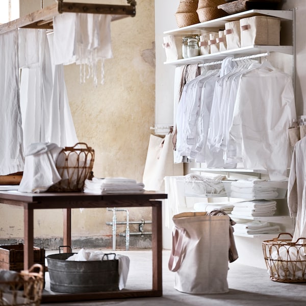 A white BOAXEL wardrobe combination with clothes on white hangers and storage baskets on top, by a brown stained table.