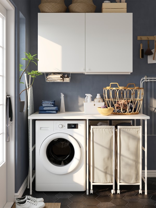 A white ENHET storage combination with two laundry bags on castors and two white wall cabinets in a blue laundry room.