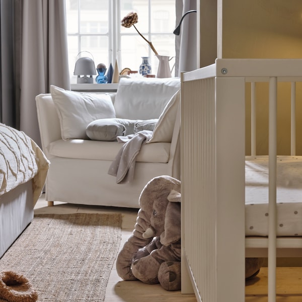 A white GULLIVER cot with an LENAST sheet on the mattress and a soft toy elephant and white BACKSÄLEN armchair behind it.