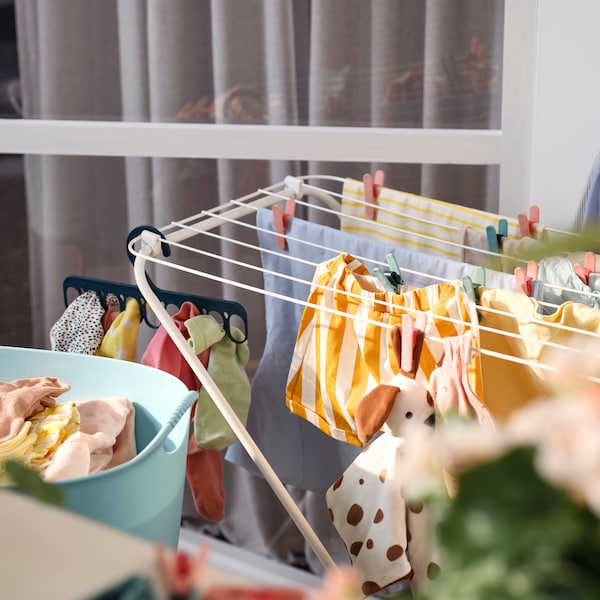 A white JÄLL drying rack with clothes held by clothes pegs and a hanger with 8 grip clips, by a flexible laundry basket.