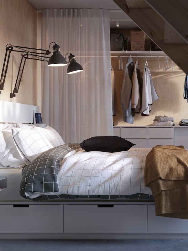 A white NORDLI bed with storage and headboard covered in VITKLÖVER bed linen, in front of an ELVARLI storage combination.
