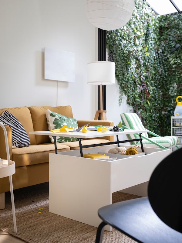 A white ÖSTAVALL adjustable coffee table with its top raised up stands in front of a yellow-brown PÄRUP sofa.
