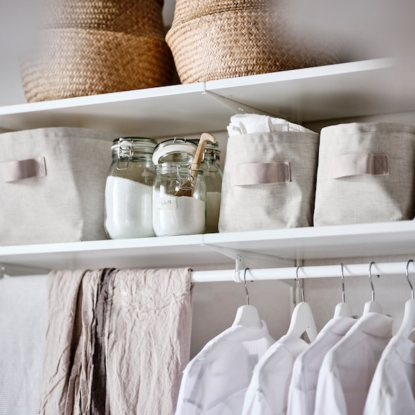 A white wardrobe combination holding seagrass baskets, beige textile storage baskets and clothes on white hangers.