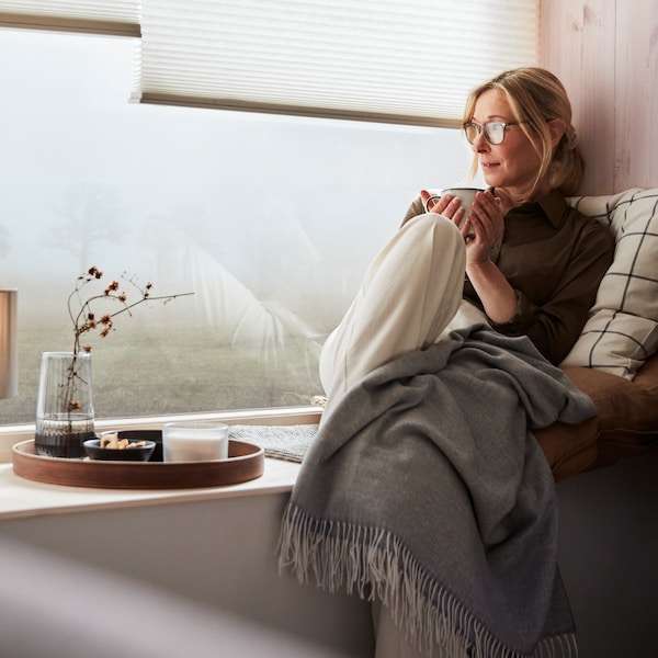 A woman in a window seat enjoying a break, with a HOLMVI throw and leaning into a cushion with a SPIKKLUBBA​ cushion cover.