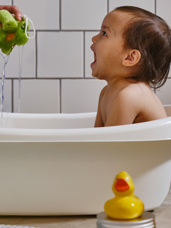 A young child sat in a white LÄTTSAM baby bath is laughing as an adult’s hand squeezes water from a green sponge.