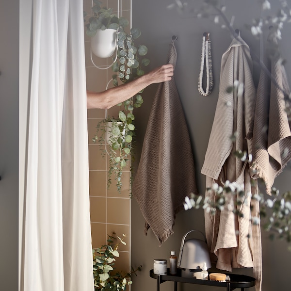 An arm reaching out of a shower for a light grey/brown towel on a hook, plus a shower curtain and artificial potted plants.