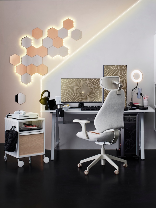 An IKEA GRUPPSPEL gaming chair in light grey, in front of a white gaming desk with two screens, tech accessories, a small storage unit on castors.